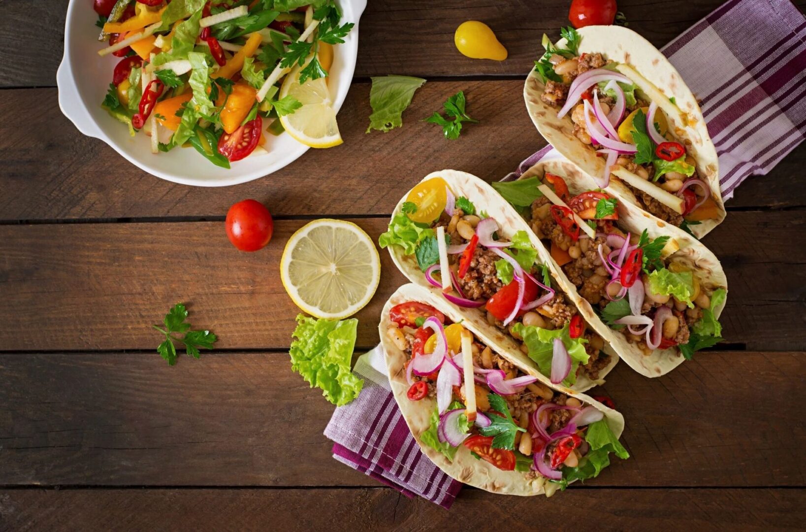 A wooden table topped with three tacos next to a bowl of salad.